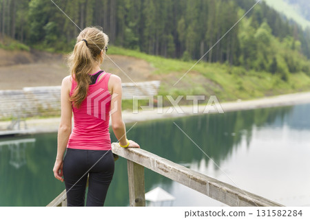 Back view of a young sportive woman in fitness clothes standing outdoors near big lake in mountains enjoying nature view. Back view of a young sportive woman in fitness clothes standing outdoors near big lake in mountains enjoying nature view. 131582284