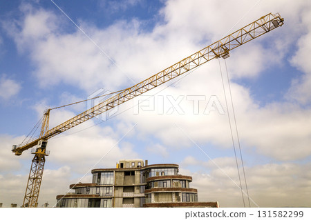 Apartment or office tall building under construction. Brick walls, glass windows, scaffolding and concrete support pillars. Tower crane on bright blue sky copy space background. Apartment or office tall building under construction. Brick walls, glass windows, scaffolding and concrete support pillars. Tower crane on bright blue sky copy space background. 131582299