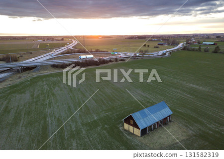 Aerial view of wooden barn in green field on background of modern highway road intersection at dawn on sunny spring day. Drone photography. Aerial view of wooden barn in green field on background of modern highway road intersection at dawn on sunny spring day. Drone photography. 131582319