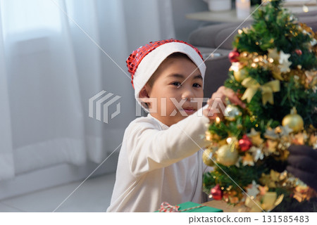 Christmas family at home, child with parent decorating holiday tree, gift here, young child wears santa hat, carefully places golden ornaments, creating warm Christmas memories. 131585483