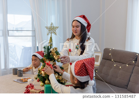 Christmas family home, parent guides child decorating holiday Christmas tree, young girl placing ornaments, boy enjoys meal, gifts are ready for celebration. 131585487