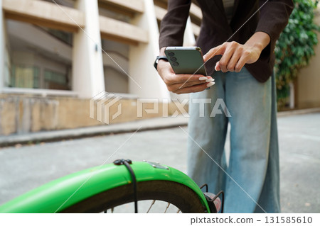 Mobile Productivity and Urban Living. A woman multitasks as she checks her phone while resting on her bicycle in a vibrant city. 131585610