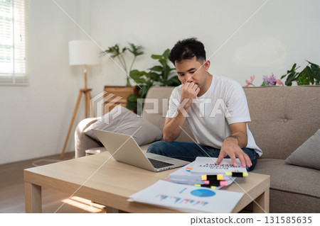 Remote Work and Focus. A man working from home, thoughtfully assessing information on his laptop and papers. 131585635