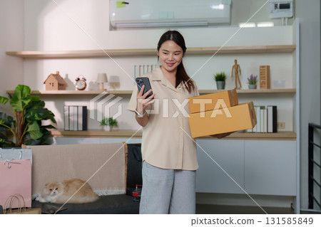 Home Shopping Delight. Woman carrying boxes while checking her phone with her cat nearby. Home Shopping Delight. Woman carrying boxes while checking her phone with her cat nearby. 131585849