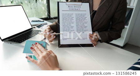 Real estate agent presenting contract to client with laptop and model houses on the table. 131586245