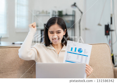 Motivated young woman celebrating success while reviewing financial reports on a video call. Motivated young woman celebrating success while reviewing financial reports on a video call. 131586342