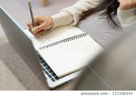 Woman taking notes while on a phone call in a comfortable home office, laptop nearby. Woman taking notes while on a phone call in a comfortable home office, laptop nearby. 131586343