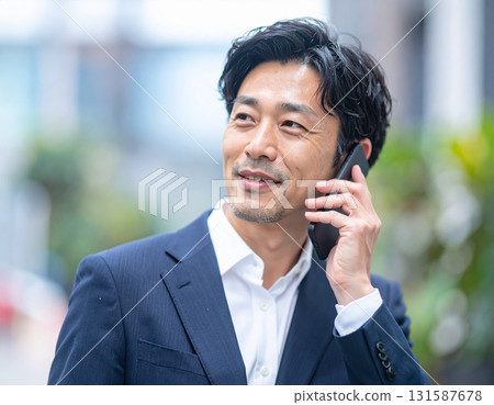 A Japanese middle-aged businessman in his 40s talking outdoors with a smartphone to his ear and a smile 131587678