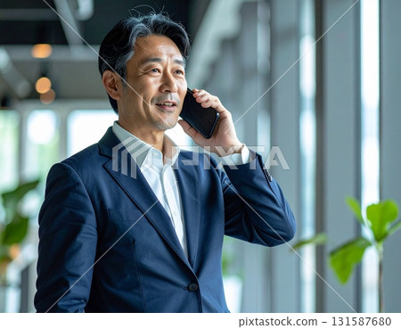 A middle-aged Japanese businessman in his 50s holds a smartphone to his ear in the office and talks on the phone with a serious expression 131587680