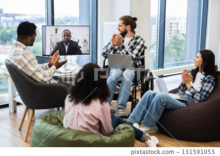 Diverse group of colleagues including man in wheelchair and a woman engaging in virtual meeting with African male speaker on screen, demonstrating teamwork, inclusivity, online collaboration. 131591053