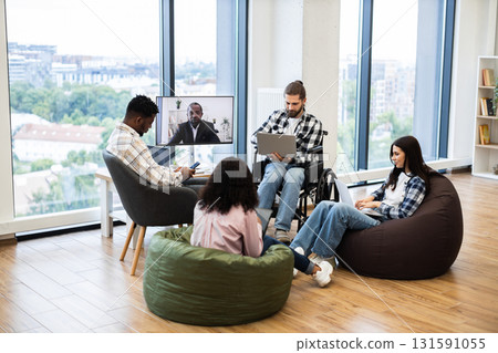 Group of multiracial coworkers, including disabled male, engaged in team meeting using video conference with male manager displayed on screen, embracing modern teamwork solutions in a casual office. Group of multiracial coworkers, including disabled male, engaged in team meeting using video conference with male manager displayed on screen, embracing modern teamwork solutions in a casual office. 131591055