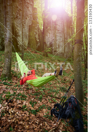 Person relaxing in bright green hammock tied between two trees in lush, green forest near rocks. Man tourist holds drink, enjoying peaceful moment surrounded by nature. Backpack and gear rest nearby. 131593430