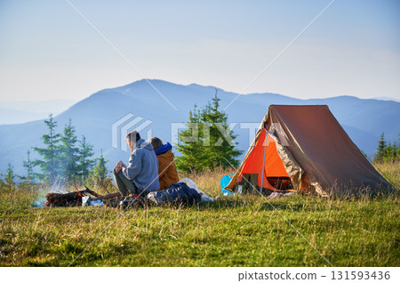 Scenic mountain campsite with two tents in morning. Back view of two campers enjoying serene view of rolling mountains. Picturesque sunrise landscape, creating tranquil and cozy outdoor experience. 131593436