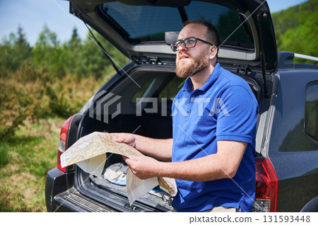 Man tourist leans on open trunk of SUV, deeply focused on map. Traveler wears blue shirt and beige pants, standing in lush, green area, carefully planning or navigating his route. 131593448