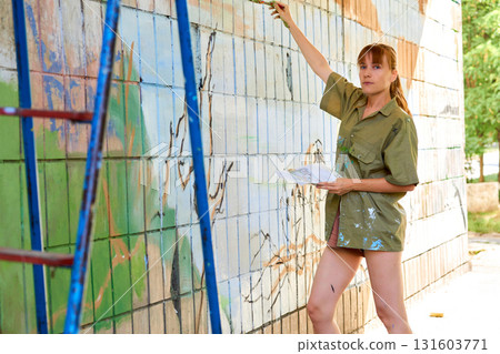 Cute graffiti artist painter with a brush, stepladder paints a wall 131603771