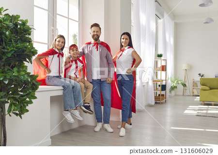 Happy family in superhero costumes standing near the windowsill at home and looking at camera. 131606029