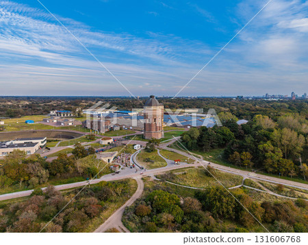 Aerial view of historical water tower amid utility buildings, green paths, and distant city skyline blending heritage architecture with infrastructure and natural surroundings. Aerial view of historical water tower amid utility buildings, green paths, and distant city skyline blending heritage architecture with infrastructure and natural surroundings. 131606768