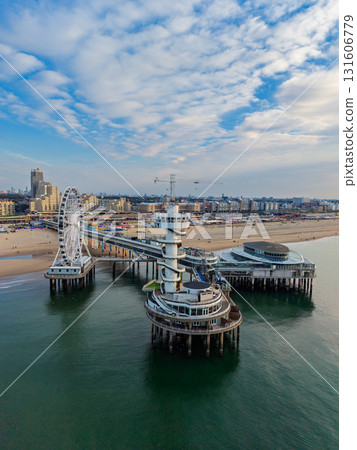 Aerial view of coastal pier with spiral observation tower, Ferris wheel, and dome structure framed by sandy beach, urban buildings, and calm sea under partly cloudy skies. 131606779