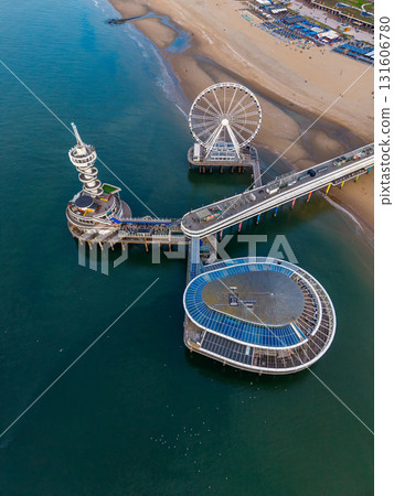 Aerial view of ocean pier with Ferris wheel, glass roofed building, and spiral tower set against busy beach and deep blue water, highlighting vibrant coastal recreation. Aerial view of ocean pier with Ferris wheel, glass roofed building, and spiral tower set against busy beach and deep blue water, highlighting vibrant coastal recreation. 131606780