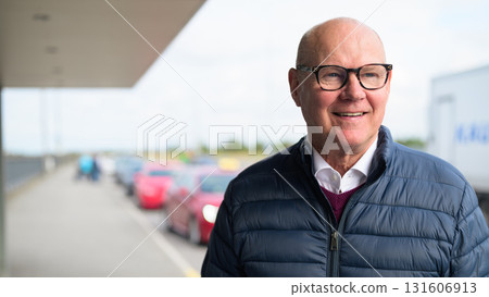 Happy senior Scandinavian man standing outdoors in front of a car park at an airport taxi area Happy senior Scandinavian man standing outdoors in front of a car park at an airport taxi area 131606913