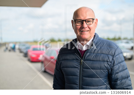 Happy senior Scandinavian man standing outdoors in front of a car park at an airport taxi area Happy senior Scandinavian man standing outdoors in front of a car park at an airport taxi area 131606914