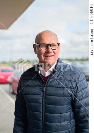 Happy senior Scandinavian man standing outdoors in front of a car park at an airport taxi area Happy senior Scandinavian man standing outdoors in front of a car park at an airport taxi area 131606916