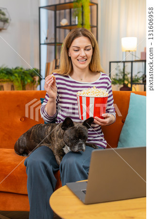 Middle-aged woman with dog laughs eats popcorn and watches favorite series or movie on laptop calm 131606958