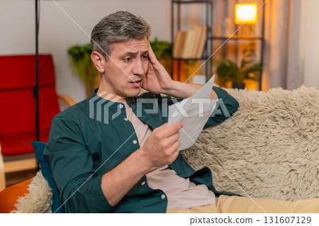 Young man reacts with stress and worry after opening envelope with credit bill at home indoors Young man reacts with stress and worry after opening envelope with credit bill at home indoors 131607129