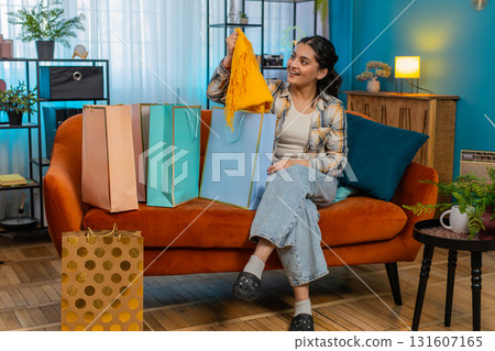 Indian woman entering room with shopping bags sitting on sofa unpacking purchases joyful excitement 131607165