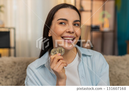 Young woman on sofa thoughtfully spinning BTC coin in hands considering crypto market investment 131607168