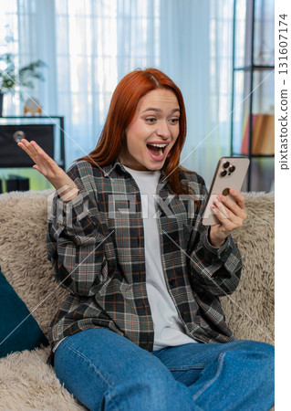 Woman sitting on sofa reading good news on smartphone raising hands up celebrating joyful success Woman sitting on sofa reading good news on smartphone raising hands up celebrating joyful success 131607174