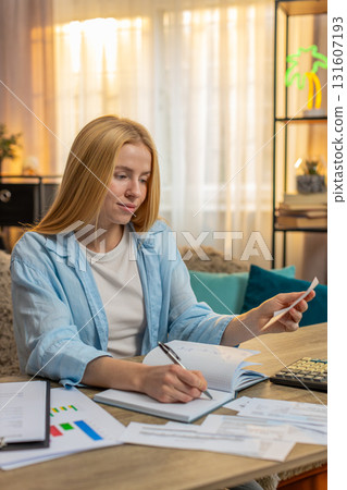 Young woman writing budget at home, table workspace, documents, calculator focused plan calm routine Young woman writing budget at home, table workspace, documents, calculator focused plan calm routine 131607193