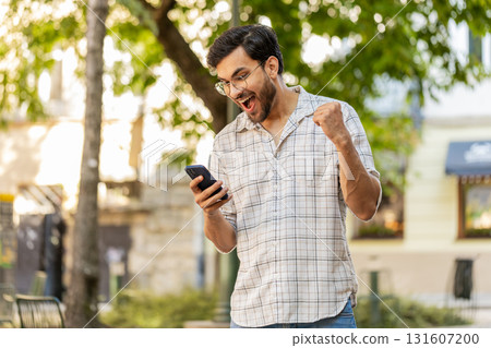 Hispanic young man using smartphone celebrating win good message news clenching fist on city street 131607200