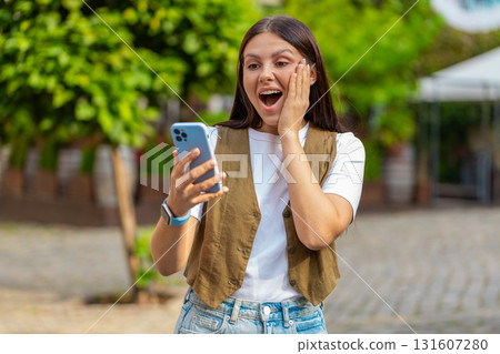 Excited European young woman using smartphone typing browsing celebrating win on urban city street Excited European young woman using smartphone typing browsing celebrating win on urban city street 131607280
