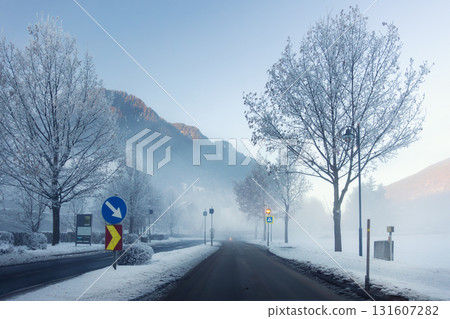 Scenic quiet winter road frosty valley covered mist fog and snow. Bare trees and distant mountains glow softly under the pale morning light. Alpine mountain village winter morning sunrise 131607282