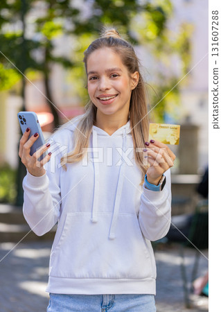 Happy young woman using credit bank card and smartphone purchases online shopping on city street Happy young woman using credit bank card and smartphone purchases online shopping on city street 131607288