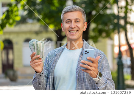 Happy rich man counting holding money dollar cash using smartphone calculator app on city street Happy rich man counting holding money dollar cash using smartphone calculator app on city street 131607289