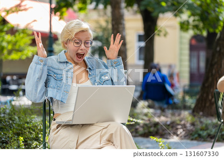 Overjoyed happy young woman working on laptop screaming in delight raises hands in city street 131607330