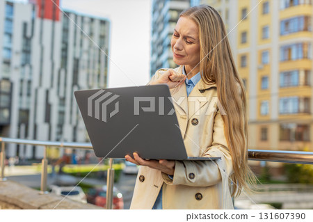Mature Caucasian woman on laptop in downtown street sees bad lottery results, looking disappointed Mature Caucasian woman on laptop in downtown street sees bad lottery results, looking disappointed 131607390