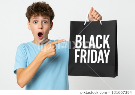 A surprised boy in a light blue shirt holding and pointing at a black shopping bag with the words Black Friday printed on it 131607394