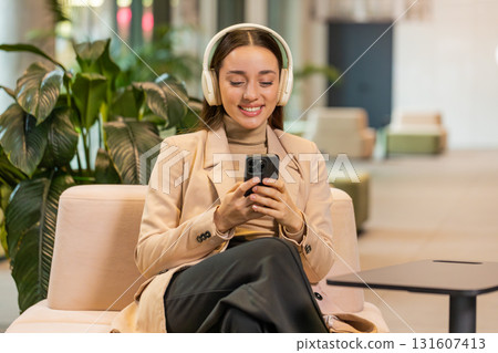 Excited young businesswoman in headphones listening energetic music on smartphone in office lobby Excited young businesswoman in headphones listening energetic music on smartphone in office lobby 131607413