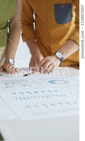 Business professionals analyzing financial reports, pointing at data points on charts and graphs lying across white desk surface Business professionals analyzing financial reports, pointing at data points on charts and graphs lying across white desk surface 131607607