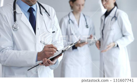 Doctor writing on a clipboard during a medical team discussion, collaborating on patient care, documentation, and healthcare strategy in a clinical setting Doctor writing on a clipboard during a medical team discussion, collaborating on patient care, documentation, and healthcare strategy in a clinical setting 131607741