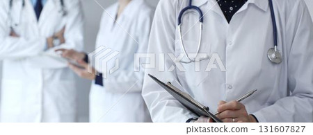 A medical professional with a stethoscope writes on a clipboard while a doctor and female assistant collaborate in a clinic, discussing patient data and healthcare information A medical professional with a stethoscope writes on a clipboard while a doctor and female assistant collaborate in a clinic, discussing patient data and healthcare information 131607827
