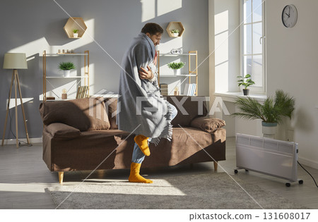 Man in blanket and socks trying to warm up in his cold house or apartment during winter Man in blanket and socks trying to warm up in his cold house or apartment during winter 131608017