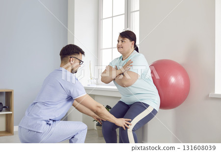 Young male nurse helping fat overweight woman doing sport exercises with fit ball. 131608039