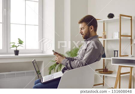 Young man with laptop using mobile phone, sitting in chair at home or at office. Young man with laptop using mobile phone, sitting in chair at home or at office. 131608048