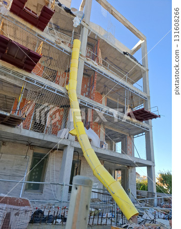 Workers are constructing a multi-story building with bright yellow debris netting and a chute attached to the roof for safely disposing of materials. Workers are constructing a multi-story building with bright yellow debris netting and a chute attached to the roof for safely disposing of materials. 131608266