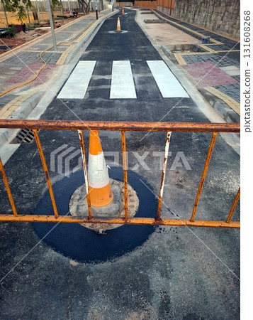 Workers are applying new road markings for pedestrian crossings on a freshly paved road in an urban area. Safety cones are positioned to guide traffic during the process. 131608268