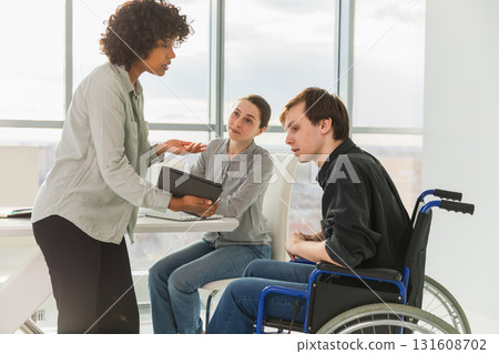Teamwork brainstorm. Diverse multiracial colleagues man in chair for people with disability working together. Group of people sitting in creative office discussing business. Young professional team Teamwork brainstorm. Diverse multiracial colleagues man in chair for people with disability working together. Group of people sitting in creative office discussing business. Young professional team 131608702